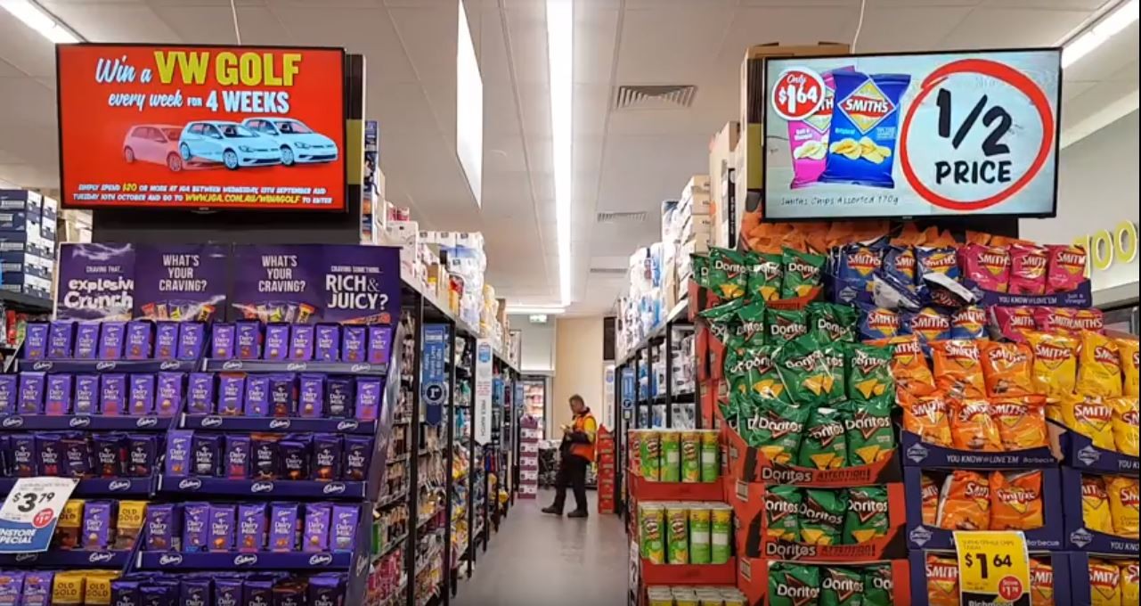 In-store ad hunt: a grocery aisle where every surface is an ad — a hanging screen promoting 'Win a VW Golf,' a '1/2 Price' Smiths chips screen, branded Cadbury and Doritos pallet displays, and shelf signage.